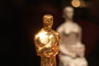 An Oscar statuette in focus with a blurred figure in the background.