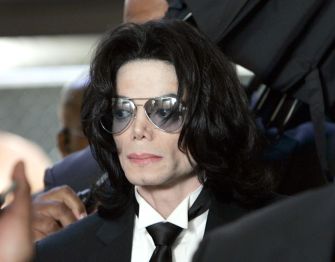 Michael Jackson wearing sunglasses and a black suit, surrounded by people during a public appearance related to his 2005 criminal trial.