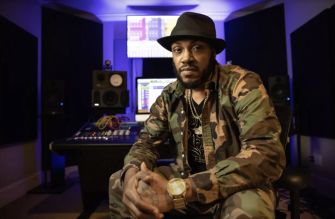 Rapper Mystikal sitting in a recording studio, wearing a camouflage jacket and hat.