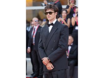 A man in formal attire wearing sunglasses stands on the red carpet at an event, with a crowd of onlookers in the background.