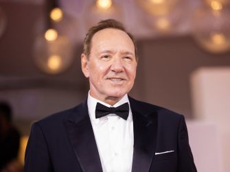 A man in a tuxedo smiles for the camera at a formal event, with blurred lights in the background.