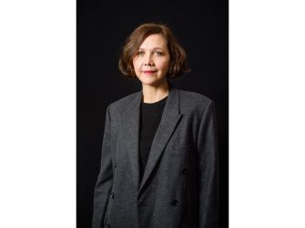A woman with shoulder-length hair wearing a grey blazer, standing in front of a dark background.