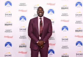 Former NFL player Marcellus Wiley smiling at an event, wearing a maroon suit with a white shirt and dark tie, standing in front of a branded backdrop.