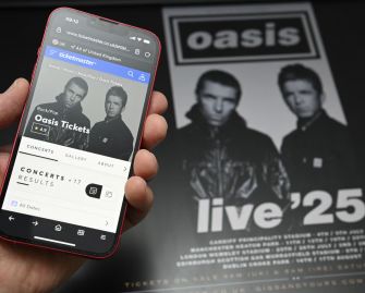 A person holding a smartphone displaying the Ticketmaster website for Oasis tickets, with an Oasis concert poster in the background.