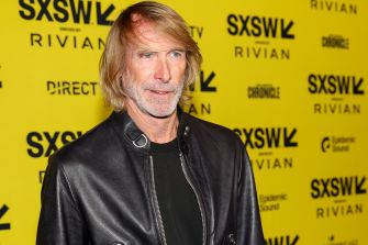 Michael Bay at a public event with a yellow backdrop displaying sponsor logos.