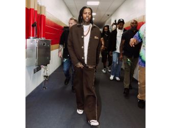 A rapper walking through a hallway, dressed in a brown outfit and flanked by several people.