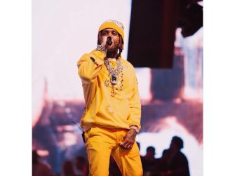A performer wearing a yellow outfit and jewelry, singing into a microphone on stage, with a blurred background of an audience and lights.