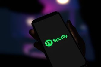A hand holding a smartphone displaying the Spotify logo against a blurred colorful background.