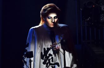 A performer in dramatic makeup and an elaborate costume, holding a microphone under spotlight.