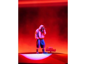 A performer on stage in a hoodie and baggy pants, illuminated by vibrant red lighting.
