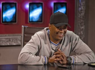 Johnny Wright, music manager, smiling while seated at a table with electronic displays in the background.