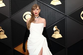 Taylor Swift on the red carpet at an awards event, wearing an elegant white dress with black gloves, posing in front of a wall featuring Grammy Awards.