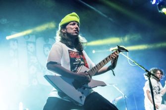 A guitarist performing on stage with a white guitar and wearing a bright green beanie, illuminated by concert lights.