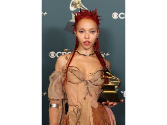 An artist holding a Grammy award while wearing an artistic outfit, posing against a dark background.