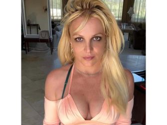 Image of a woman with long blonde hair, wearing a light-colored off-the-shoulder top, sitting indoors with sunlight coming through the windows.