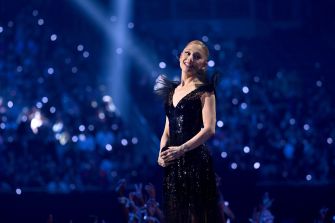Ariana Grande performing on stage in a glamorous black gown, with a vibrant crowd and lights in the background.