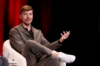 A man in casual attire speaking at an event, gesturing with his hands, against a backdrop of red and black lighting.