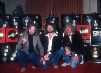 Three musicians pose in front of gold and platinum records at a celebration event.