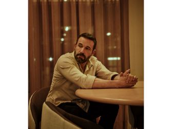 A man sitting at a table with a serious expression, surrounded by soft lighting and curtains.