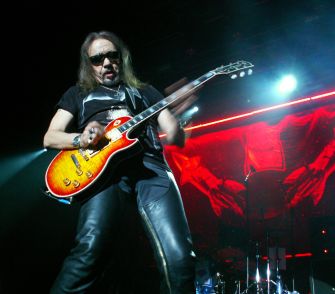 A musician performing on stage with an electric guitar, wearing sunglasses and a black outfit.
