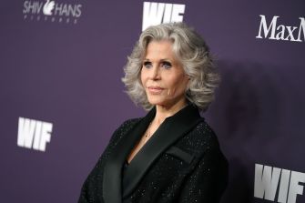 Jane Fonda at an event, wearing a stylish black outfit and posed against a purple backdrop with industry logos.
