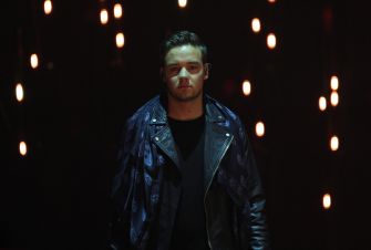 A singer performing on stage with dramatic lighting in the background.