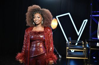 A woman with curly hair wearing a red sequined outfit stands confidently in front of a stage setup, celebrating her victory on a music competition show.