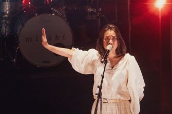 A performer on stage with a microphone, gesturing with one hand, against a backdrop featuring a drum set with the name "mitski".