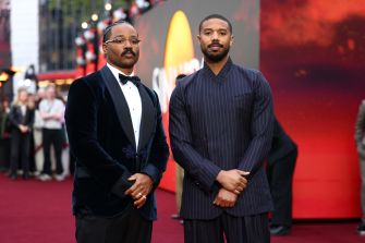 Two men in formal attire posing on the red carpet at an event, with a blurred background featuring a display related to the film "Sinners."