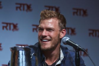 Actor Alan Ritchson at a red carpet event, posing in a dark outfit against a promotional backdrop.
