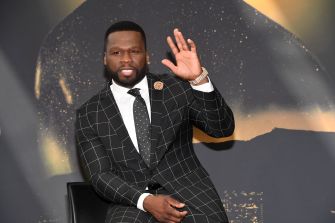 50 Cent raising his hand and smiling while wearing a stylish suit during a public appearance.