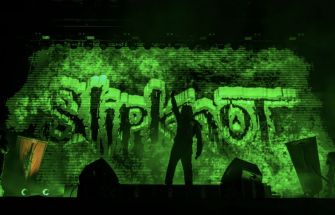 A performer silhouetted against a green backdrop featuring the Slipknot logo during a concert.
