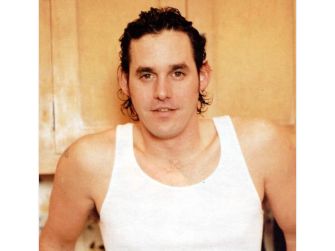 A man in a sleeveless white shirt, standing in front of a kitchen setting.