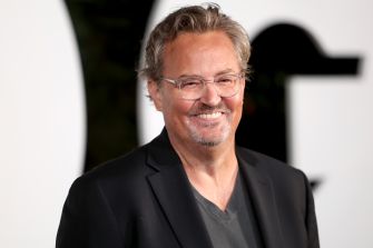 Matthew Perry smiling at a public event, wearing glasses and a black blazer over a gray shirt.