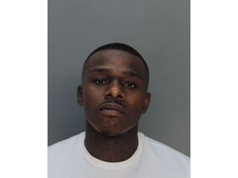 A close-up portrait of a male rapper with a serious expression, featuring short hair and visible tattoos on his neck.