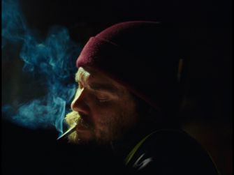 A close-up of a man with a beanie, smoking and surrounded by smoke in a dimly lit setting.