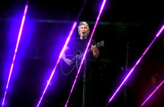 Roger Waters performing on stage with a guitar, surrounded by colorful laser lights.