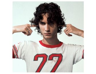 A young male artist poses with a serious expression, wearing a vintage-style t-shirt, and holding his fingers to his ears.