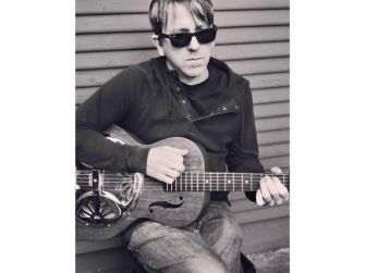 A black and white photo of Greg Brown sitting down with a guitar, wearing sunglasses and a black jacket.