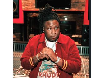 A young music producer in a recording studio, featuring red attire and dreadlocks, looking thoughtfully at the camera.
