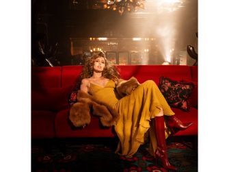 A woman wearing a yellow dress and red boots sits on a red couch in a dimly lit room, exuding a glamorous and confident vibe.
