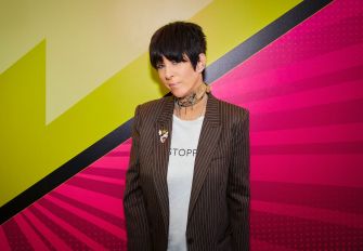 Diane Warren wearing a striped blazer and t-shirt, standing in front of a vibrant, colorful background.