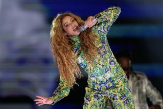 Shakira performing on stage at a concert, dressed in a colorful, patterned outfit while singing into a microphone.
