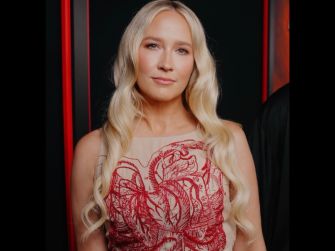 Actor Anna Camp posing in an elegant red dress with long, wavy hair, against a dark background with red accents.