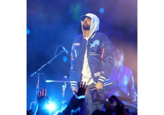 Eminem performing on stage, wearing a blue jacket and a hoodie, with a vibrant light backdrop.