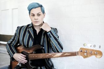 Musician Jack White poses with a guitar, wearing a striped suit and styled hair, in a modern setting.