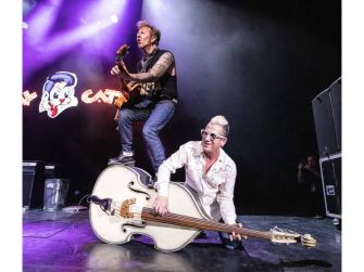 Two musicians performing energetically on stage, one playing guitar while standing on a large upright bass.