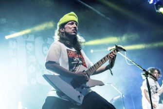 A guitarist performing on stage with a guitar and wearing a vibrant beanie, set against dynamic stage lighting.