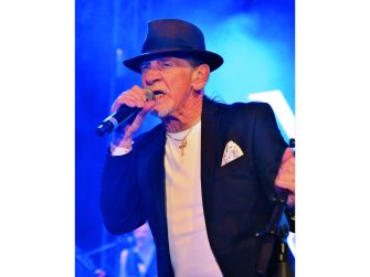Alex Ligertwood performing on stage, wearing a hat and holding a microphone, with blue lighting in the background.