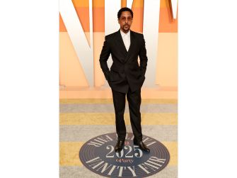 A man in a formal black suit stands confidently at an event with a colorful backdrop and a circular logo on the floor.